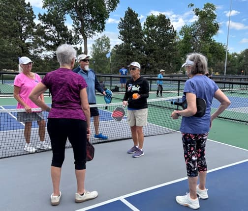 An instructor holds up a pickleball while explaining a shot to four players at the net at Monument Valley Park